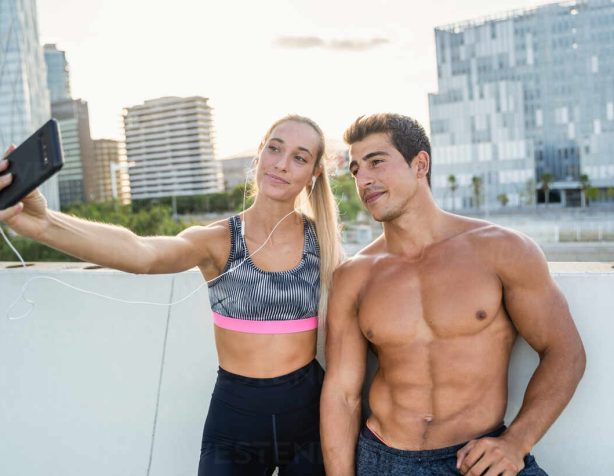 Young slim woman n sportswear and strong muscular shirtless man taking selfie on smartphone after fitness workout together in city